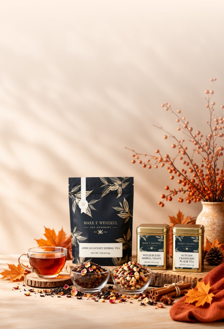 Mark Windell tea products displayed on a warm beige surface with autumn props, including a black pouch of Darjeeling Sumur Himalayan Tea, two gold tins of Masala Chai Mix, loose tea blends in small glass bowls, a cup of amber tea, cinnamon sticks, and dried orange berries in a vase.